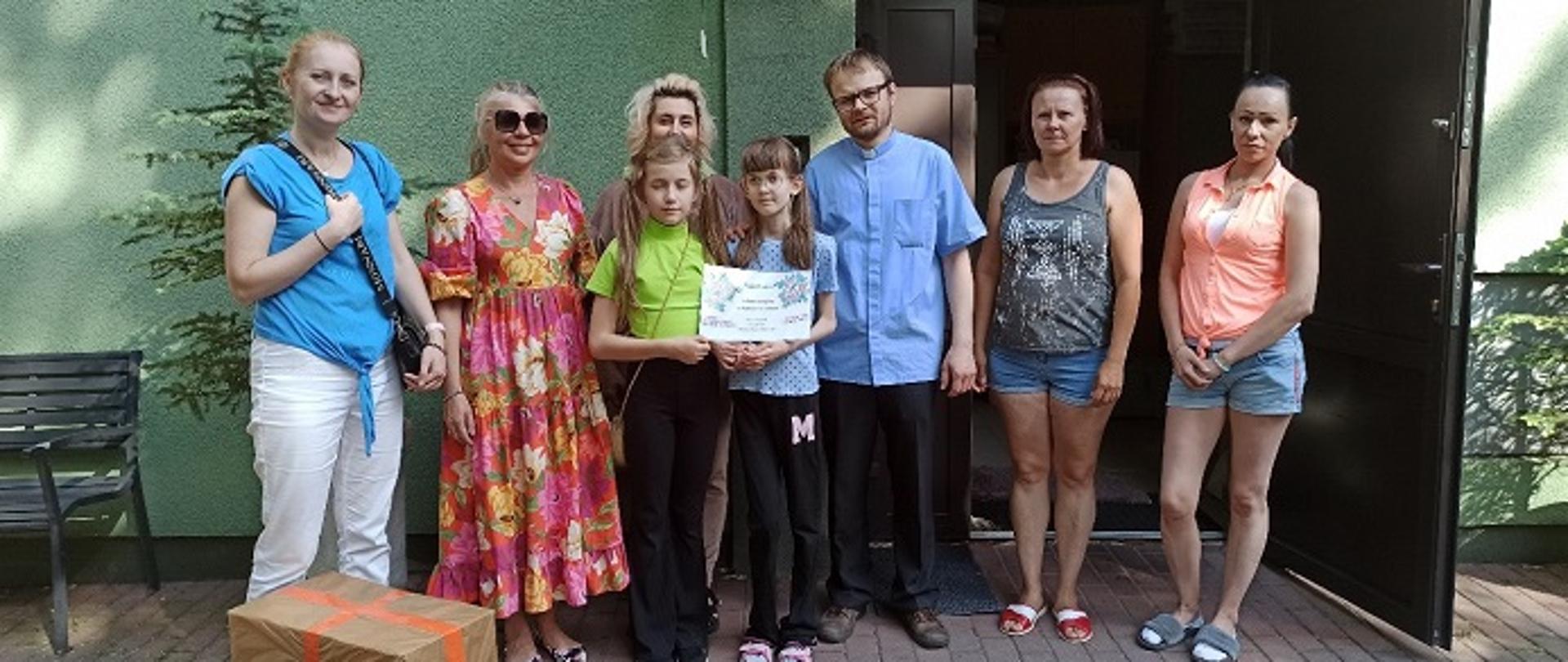 Przed budynkiem Domu Samotnej Matki stoją dwie uczennice z księdzem, rodzicem oraz mieszkankami placówki. 