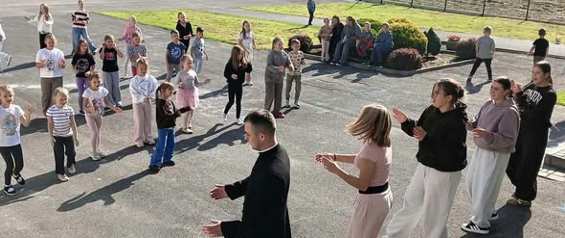 Na zdjęciu widać grupę dzieci i młodzieży uczestniczących w zajęciach na świeżym powietrzu. Na pierwszym planie znajduje się ksiądz ubrany w czarną sutannę, który wykonuje gesty, a obok niego kilka kobiet i dziewcząt naśladuje jego ruchy. Przed nimi stoją dzieci ustawione w luźnym kręgu, które również biorą udział w aktywności.
Wydarzenie odbywa się na terenie szkolnym lub rekreacyjnym, na asfaltowym placu, w słoneczny dzień. W tle widoczne są boisko do koszykówki, ogrodzenie, drzewa i zielone tereny. Słońce rzuca długie cienie na ziemię, tworząc ciepłą, jesienną atmosferę.
Zdjęcie ukazuje wspólną aktywność, integrację i radosne spędzanie czasu w grupie. ☀️