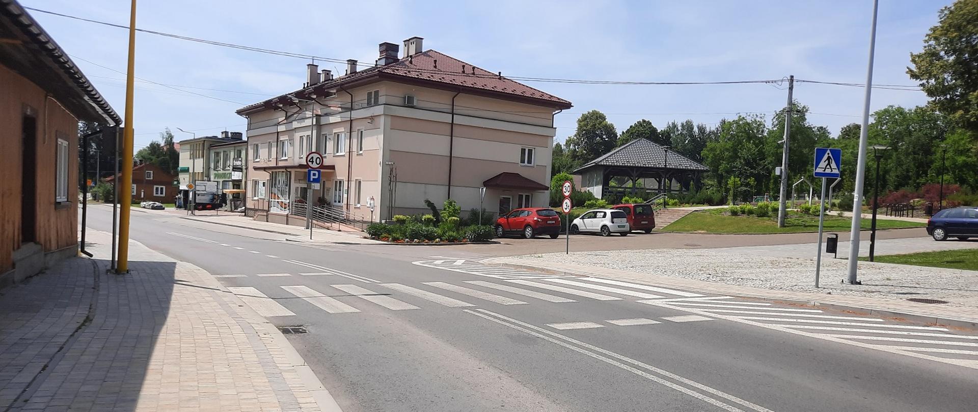 przejście dla pieszych obok dużego jasnego budynku, w tle zaparkowane samochody, słoneczna pogoda, 