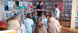 Zdjęcie przedstawia dzieci siedzące na ławkach w bibliotece, uczestniczące w zajęciach z przypasowywania elementów książek do odpowiednich gatunków. Elementy na kartonikach podaje prowadząca zajęcia a dzieci wkładają je do kieszonek na plakatach zawieszonych na regałach.