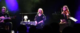 Trzy osoby siedzące na scenie, Stanisława Celińska oraz dwoje muzyków, kobieta grająca na skrzypcach i mężczyzna grający na gitarze. Przy Stanisławie stolik z lampką. Zdjęcie zrobione w trakcie koncertu.