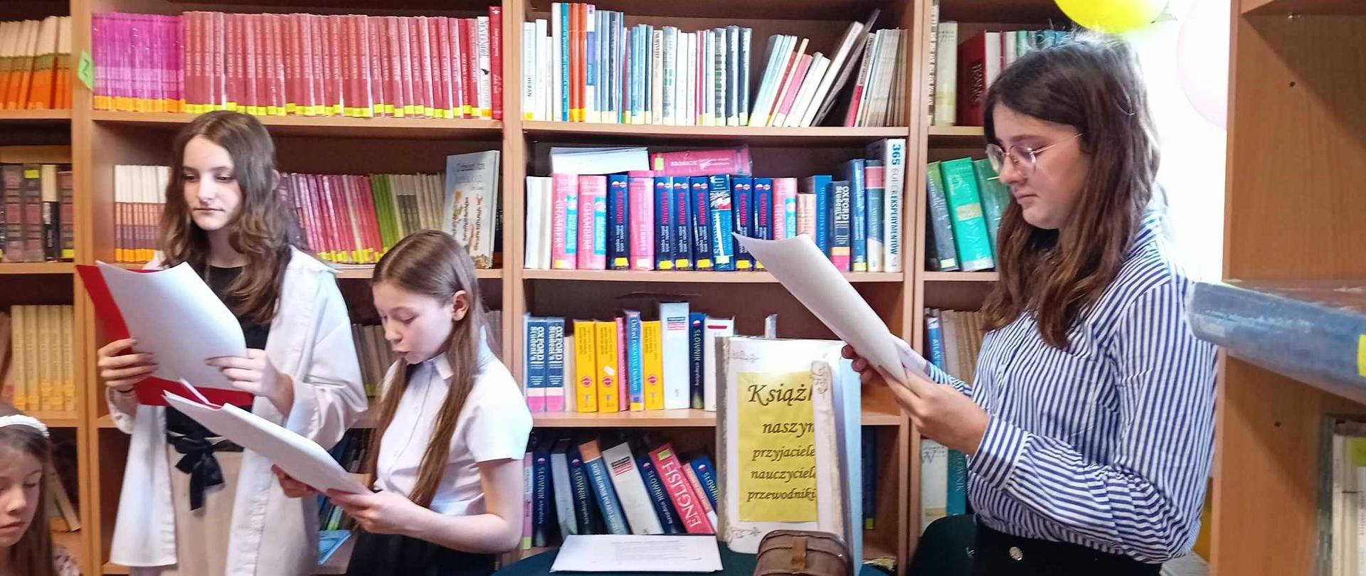 Trzy dziewczyny czytają z kartek trzymanych przed sobą. Przy ścianie stoi wysoki regał z książkami.