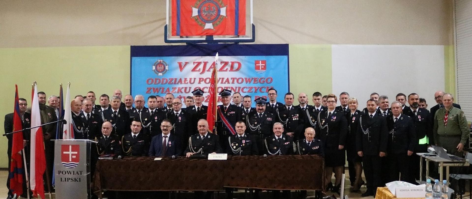 Uczestnicy zjazdu w sali gimnastycznej Zespołu Szkół Ogólnokształcących i Technicznych w Lipsku. Na pierwszym planie stoi mównica z napisem Powiat lipski i herbem Powiatu. W tle tablica z nazwa zjazdu oraz flaga straży.