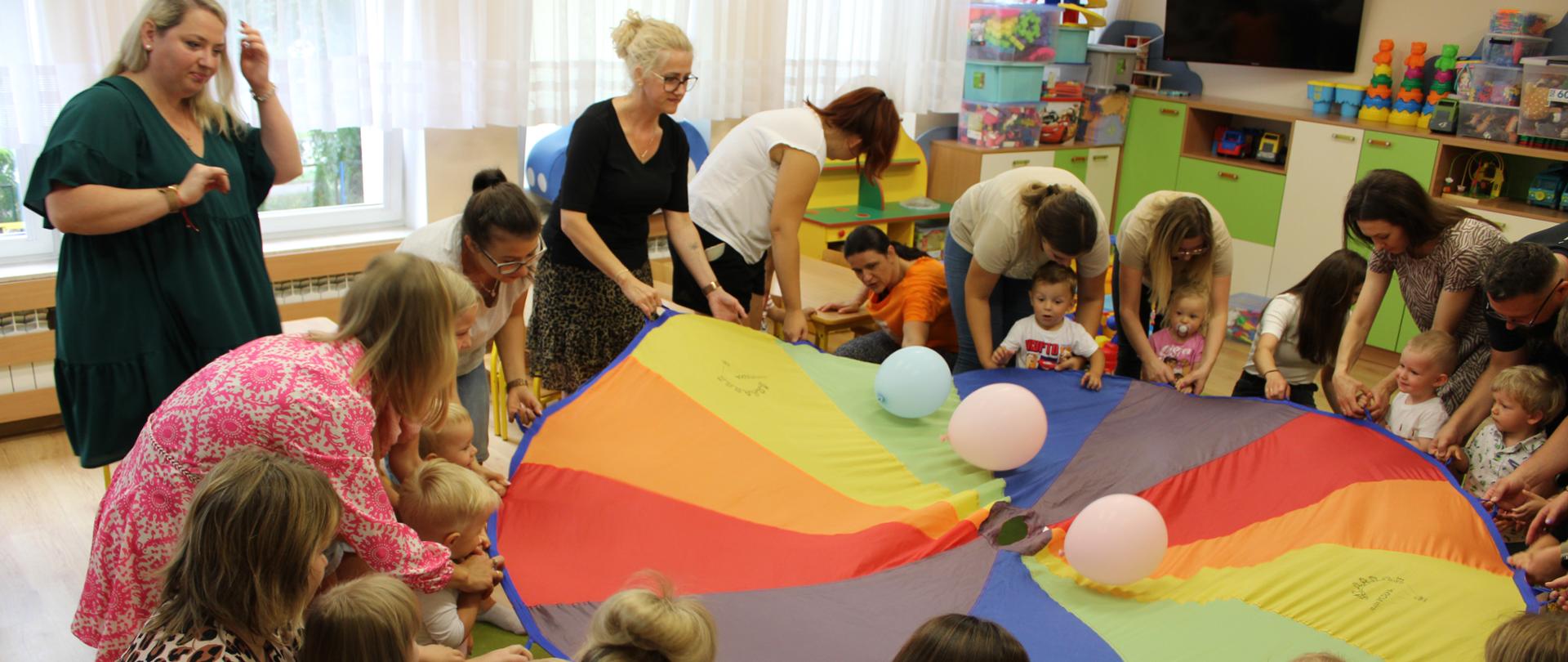 Na zdjęciu widać kolorową chustę animacyjną, a w koło niej rodziców z dziećmi.