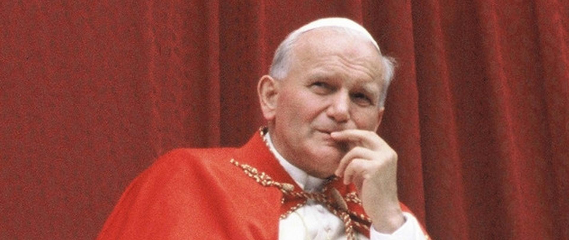 Papież Jak Paweł II w białej sutannie z czerwoną mozzetą (pelerynką) na ramionach, zamyślony, z dłonią uniesioną do twarzy, spogląda w dal; za nim bordowa pofalowana tkanina