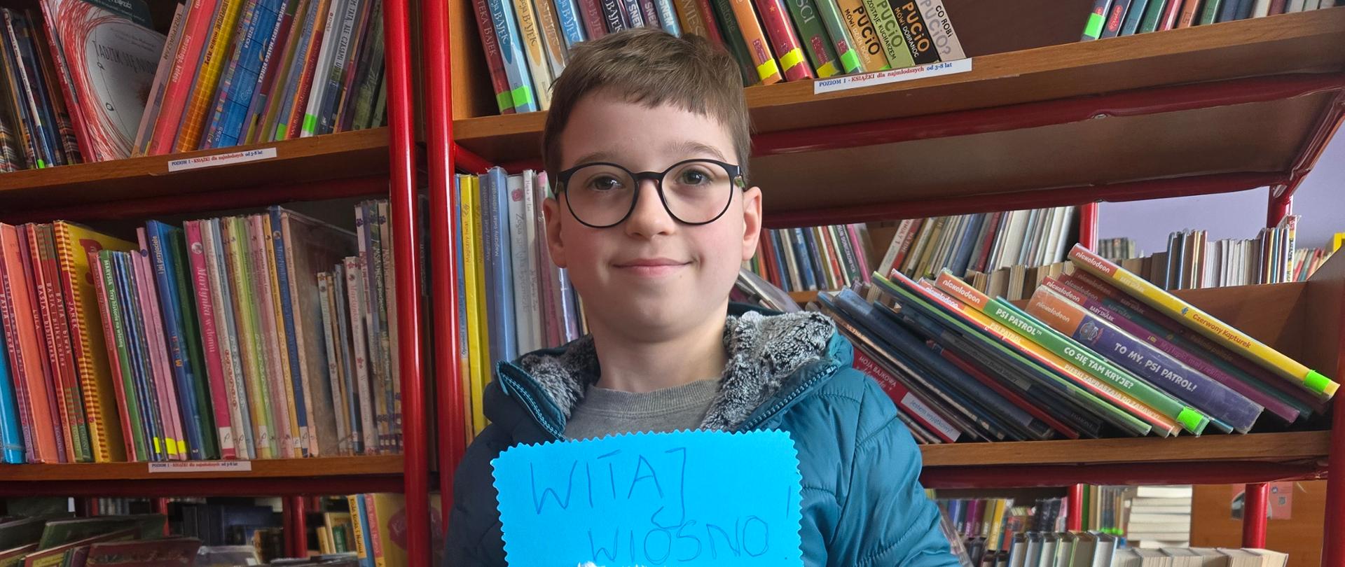 Uczeń na tle regału z książkami prezentuje swoją pracę