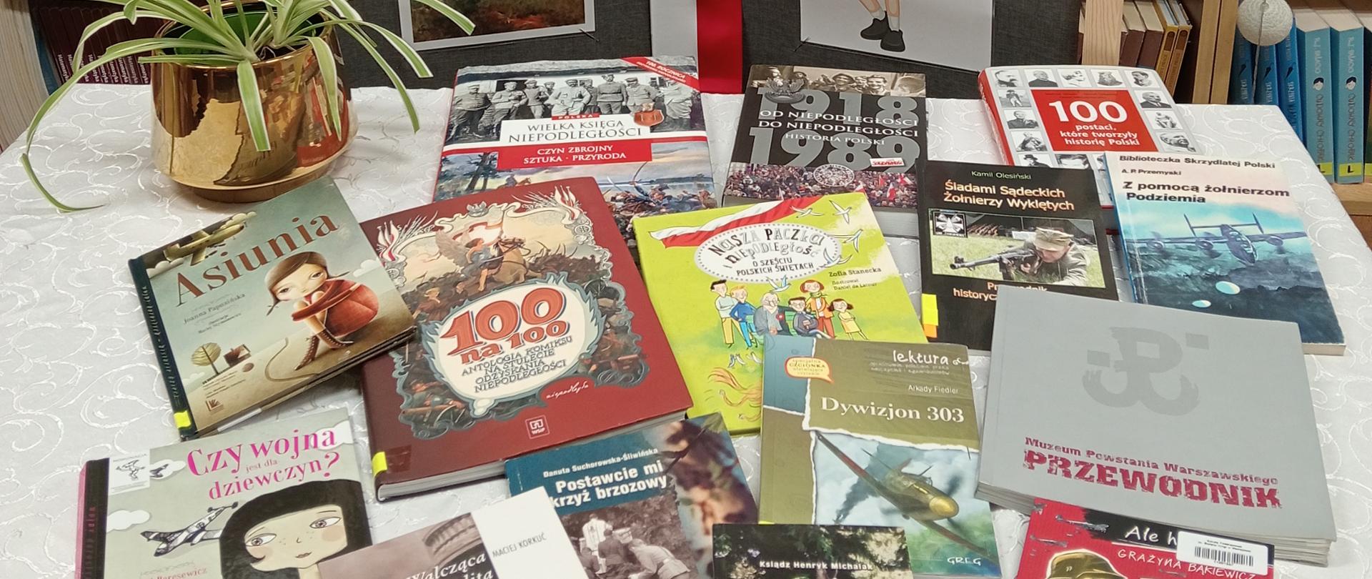 W bibliotece na stole przykrytym białym obrusem leżą książki o tematyce niepodległości i wojny, obok kwiat w złotej doniczce i gazetka z symbolami niepodległości
