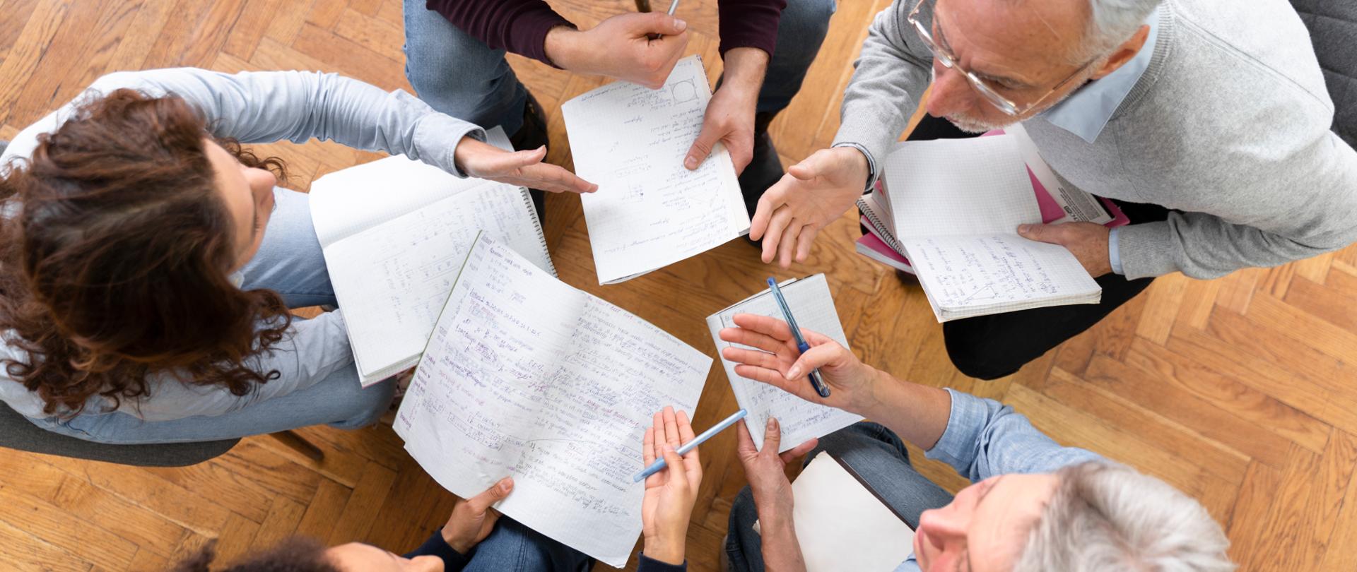 Grupa osób siedzących w kręgu z notesami i długopisami, wspólnie omawiająca notatki i rozwiązania. Widok z góry na spotkanie edukacyjne lub warsztaty.