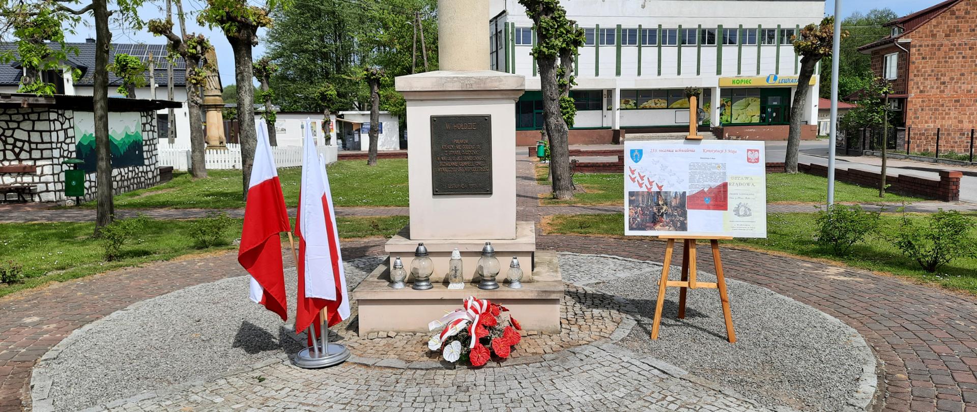 Zdjęcie przedstawia Pomnik Wolności w parku w Wielgomłynach. Pod pomnikiem kwiaty i znicze. Z lewej strony w stojaku 3 flagi biało-czerwone, z prawej sztaluga z rysem historycznym uchwalenia Konstytucji 3 Maja.