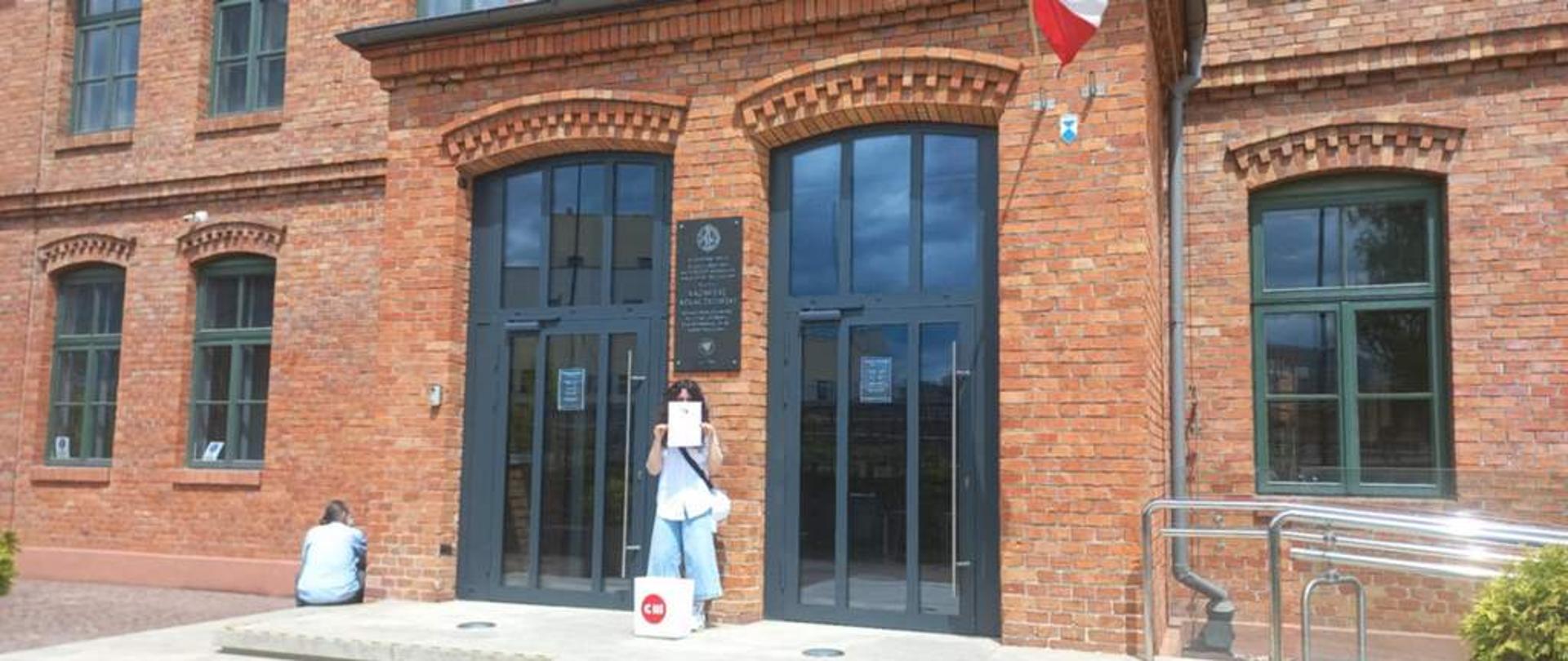 Budynek z czerwonej cegły, prawdopodobnie zabytkowy, z dużymi oknami i drzwiami w ciemnych ramach. Przed jednym z wejść stoi uczennica, trzymająca w dłoniach dyplom, częściowo zasłaniając nim twarz. Na ziemi obok niej leży biała torba z czerwonym logo. Na fasadzie budynku, między drzwiami, wisi tablica. W lewym dolnym rogu zdjęcia widać siedzącą osobę, a w prawym górnym rogu fragment flagi.