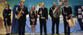 Na zdjęciu widnieje Orkiestra OSP Sokołów Podlaski. Członkowie orkiestry ubrani są w ciemne mundury. W rękach trzymają instrumenty dęte. W tle na błękitnym materiale widnieje dekoracja o tematyce muzycznej.