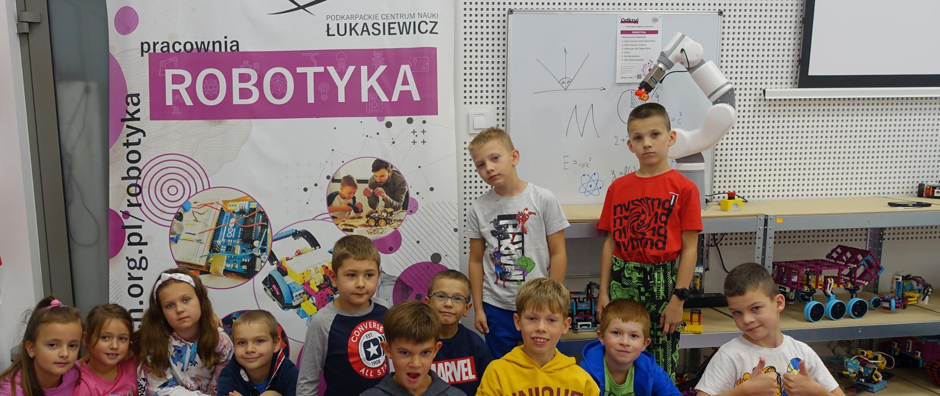 Uczniowie na tle plakatu pracowni robotyki w PCN Łukasiewicz