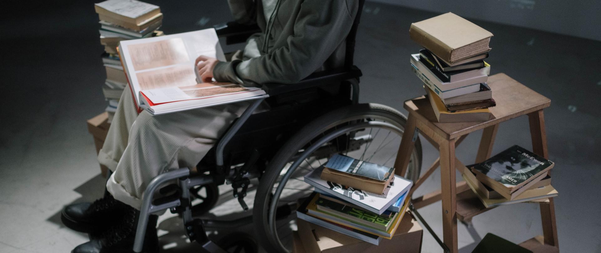 Człowiek siedzi na wózku inwalidzkim. Na kolanach trzyma dużą książkę. Z jego prawej i lewej strony stoją książki ułożone w stosy. Z lewej strony stoi mała drewniana drabinka oraz kartony. Na nich ułożone książki.