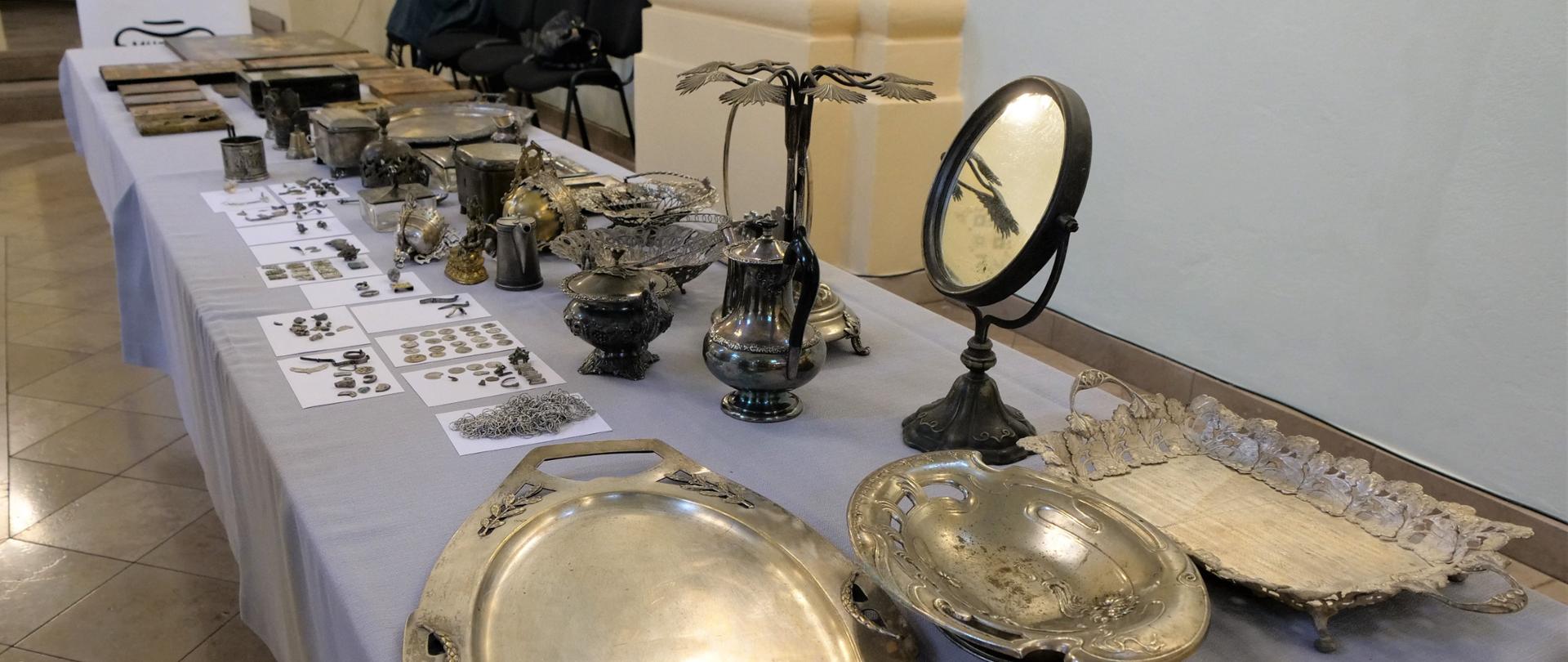 Zdjęcie przedstawia zabytkowe przedmioty ułożone na stole wewnątrz budynku. Na stole leżą złote patery, a także lustro, wazony i monety
