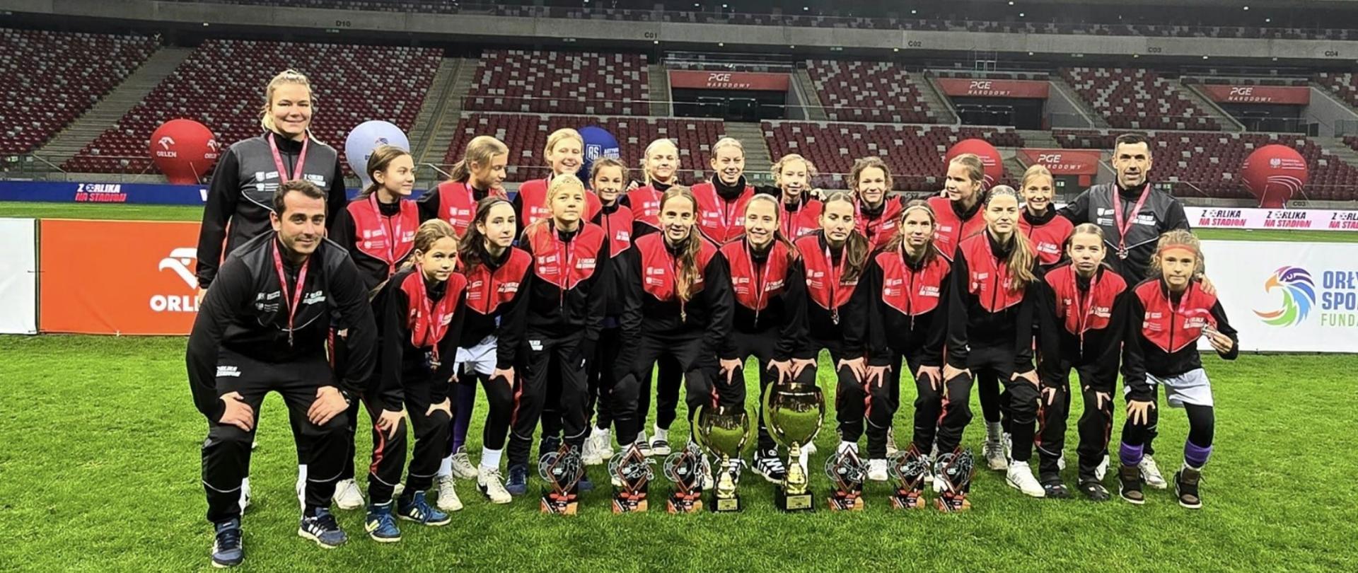Drużyna piłkarska dziewczyn w czarno czerwonych strojach wraz z trenerami pozują do zdjęcia na zielonej murawie stadionu. W tle czerwono białe trybuny.