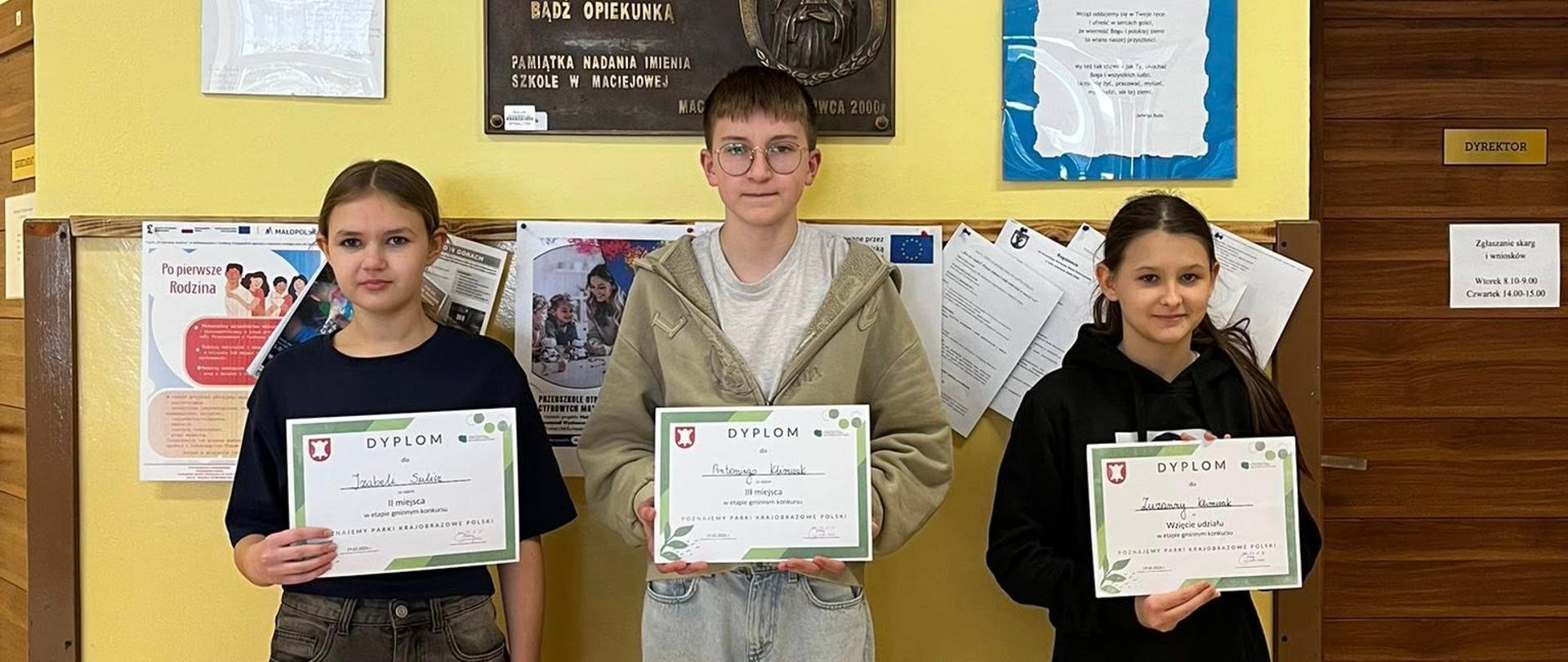Dwie uczennice i jeden uczeń stoją na korytarzu szkolnym pod obrazem patronki szkoły Świętej Kingi, trzymają w ręku dyplomy