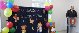 Wójt Gminy Szumowo Andrzej Pskiet stoi za przezroczystą mównicą z herbem i napisem "Gmina Szumowo. Obok niego znajduje się dekoracyjna ścianka z napisem "Tu zaczyna się przygoda...". Ścianka ozdobiona jest kolorowymi balonami, postaciami z bajek, zwierzętami oraz pluszowymi wiewiórkami.