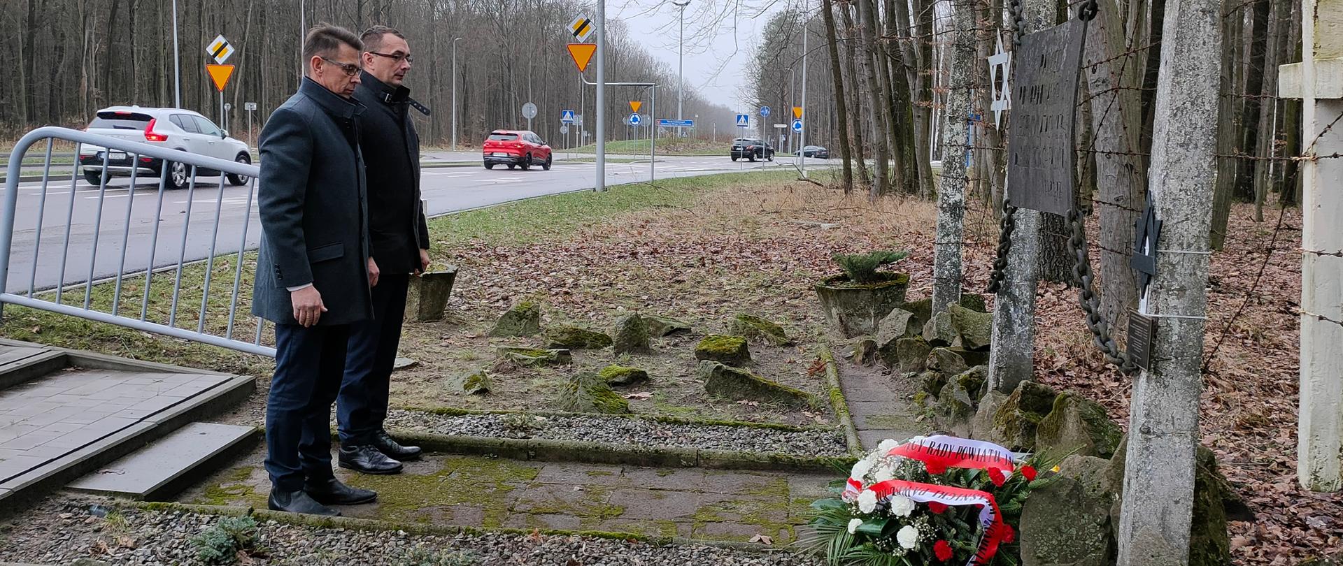 Na zdjęciu pomnik ofiar holokaustu, przy którym dwie osoby złożyły wieńce. Obóz znajduje się przy drodze asfaltowej, na której trwa ruch uliczny. Pomnik stoi na skraju lasu, tuż za nim rosną drzewa.