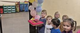 Przed wejściem do nowej sali stoją przedszkolaki, jeden chłopiec trzyma w ręku atłasową poduszkę z nożyczkami do przecięcia wstęgi inauguracyjnej, przy wejściu balony