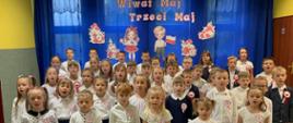 Uczniowie i dzieci z oddziału przedszkolnego ubrani na galowo w białe koszule i granatowe spodnie i spódniczki, z przypiętymi do koszulek kotylionami, śpiewają na tle szafirowej dekoracji z napisem "Wiwat Maj, Trzeci Maj"