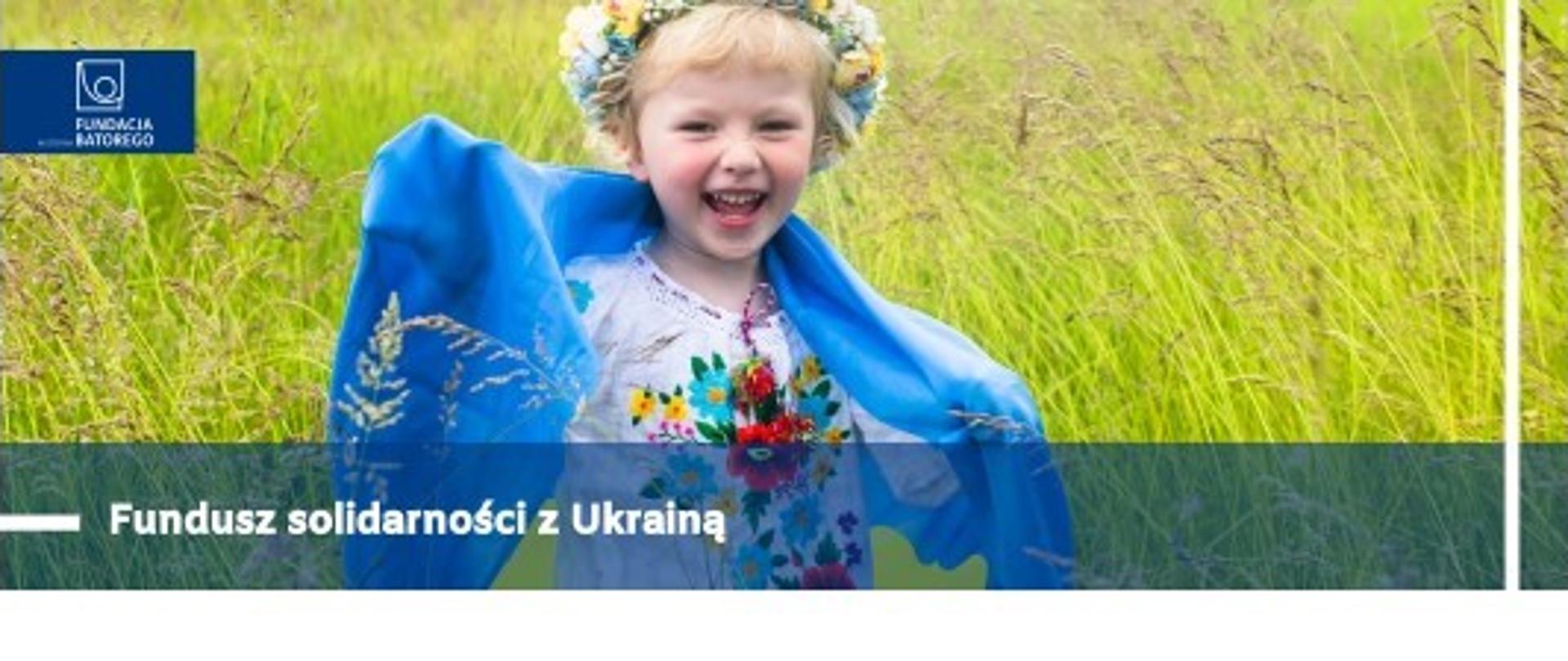 Fundusz Solidarności z Ukrainą. Fundacja Batorego. Dziewczynka w wianku z niebiesko-żółtą flagą Ukrainy. W tle bujne trawy.