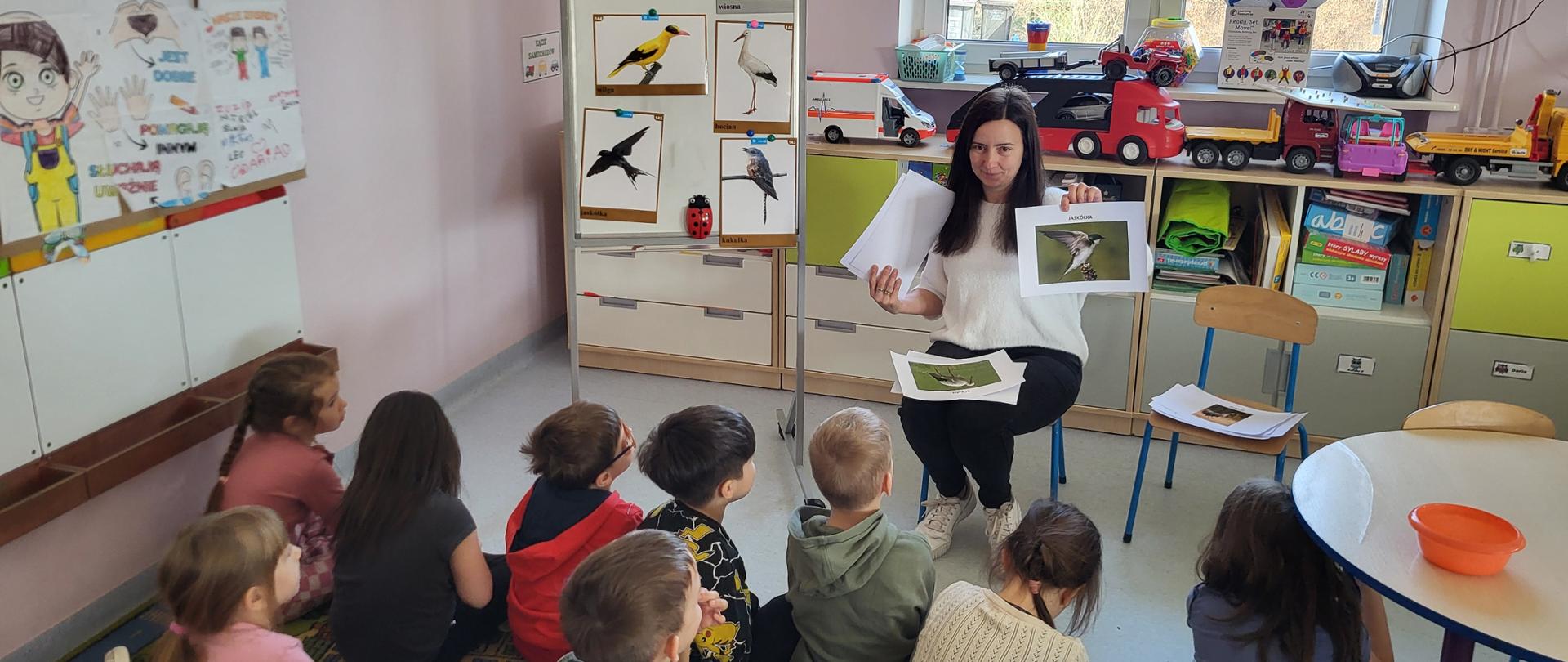 W klasie na krześle siedzi kobieta. Przed nią na dywanie siedzą dzieci. Kobieta pokazuje dzieciom zdjęcia ptaków. Za kobietą widać okno i regał z zabawkami.