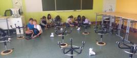 Sala lekcyjna. Pod ścianami na podłodze w 3 grupach siedzą uczniowie z tabletami. Dzieci sterują swoim robotem, który przemieszcza się pomiędzy kszesełkami, które rozstawione są na podłodze. Stoliki zsunięte są pod ścianę.