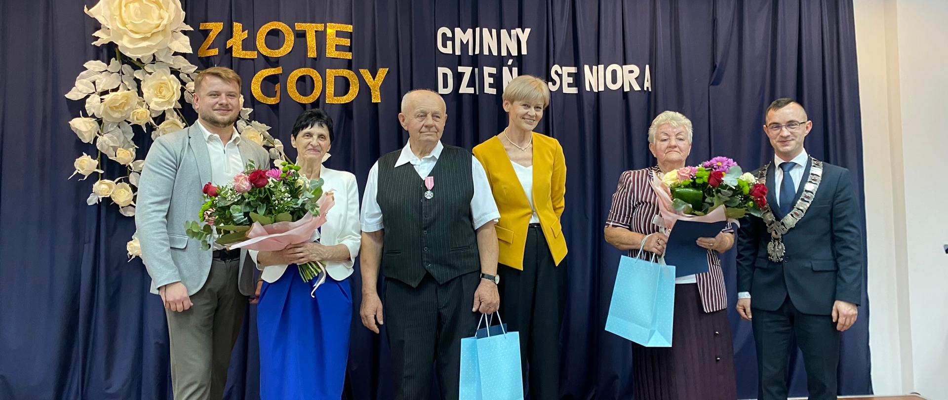 Sześć osób stoi razem na scenie podczas uroczystości Złotych Godów i Gminnego Dnia Seniora. W centrum stoją dwie nagrodzone pary seniorów. Po bokach stoją dwaj mężczyźni i kobieta w żółtej marynarce, reprezentujący władze lub organizatorów. Seniorzy trzymają bukiety kwiatów i niebieskie torby z upominkami. W tle widoczna jest ciemnoniebieska zasłona z napisem "ZŁOTE GODY GMINNY DZIEŃ SENIORA" i dekoracją z białych róż.
