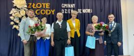 Sześć osób stoi razem na scenie podczas uroczystości Złotych Godów i Gminnego Dnia Seniora. W centrum stoją dwie nagrodzone pary seniorów. Po bokach stoją dwaj mężczyźni i kobieta w żółtej marynarce, reprezentujący władze lub organizatorów. Seniorzy trzymają bukiety kwiatów i niebieskie torby z upominkami. W tle widoczna jest ciemnoniebieska zasłona z napisem "ZŁOTE GODY GMINNY DZIEŃ SENIORA" i dekoracją z białych róż.