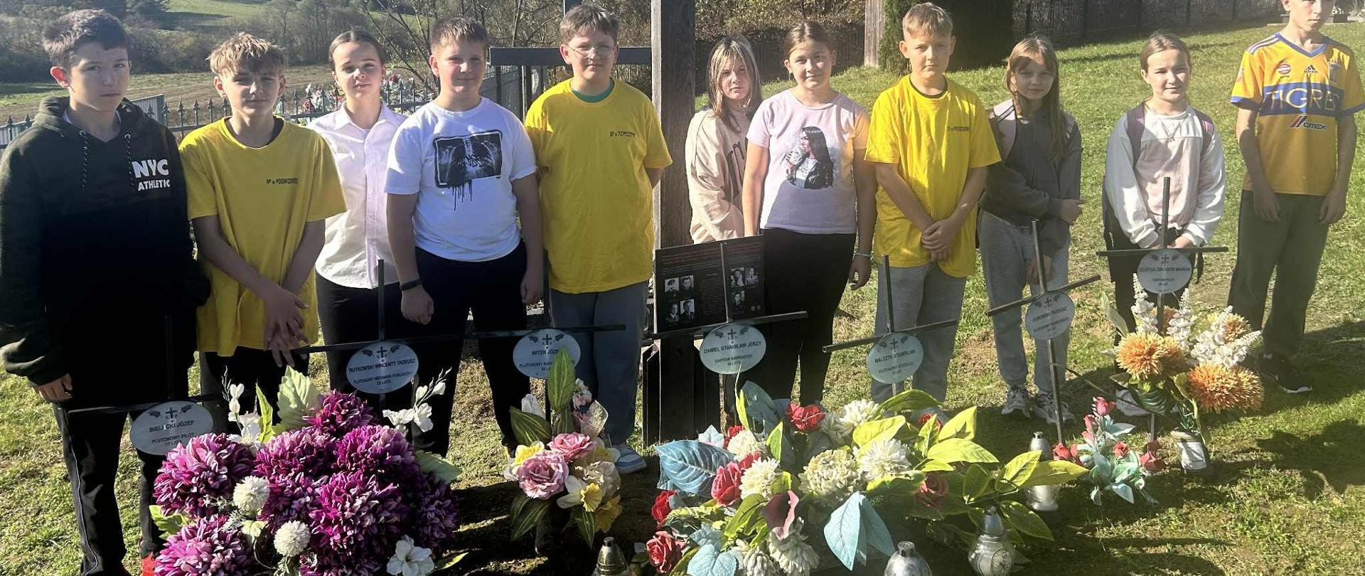 Dzieci w koszulkach koloru żółtego i z napisem wolontariat stoją na cmentarzu za grobem w którym pochowani są żołnierze. Przed nimi kwiaty i znicze.