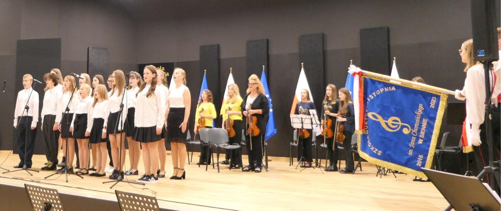 Uczniowie szkoły prezentują swoje umiejętności na scenie przy sztandarze szkoły.