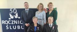 Państwo Danuta i Sławomir na tle granatowej tablicy z napisem wraz z Wójtem, Z-cą Wójta i Z-cą Kierownika USC