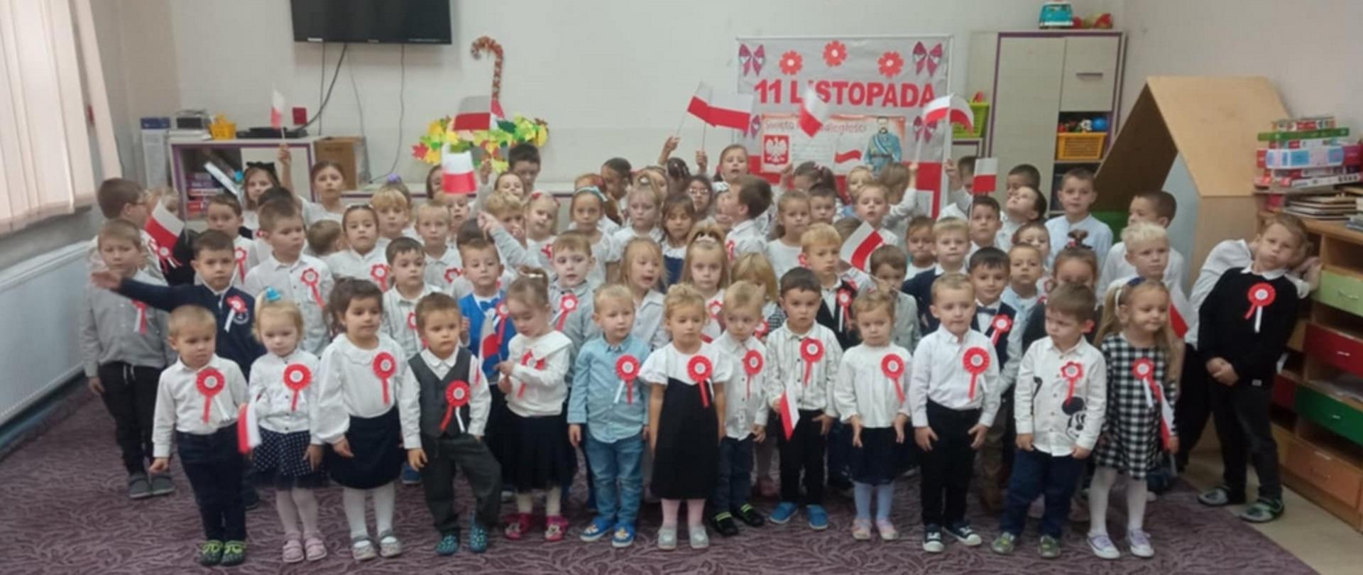 W sali przedszkolnej duża grupa dzieci pozuje do zdjęcia w strojach galowych, z przypiętymi do koszul biało-czerwonymi kotylionami. Część dzieci macha biało-czerwonymi papierowymi flagami. Z tyłu napis 11 listopada.
