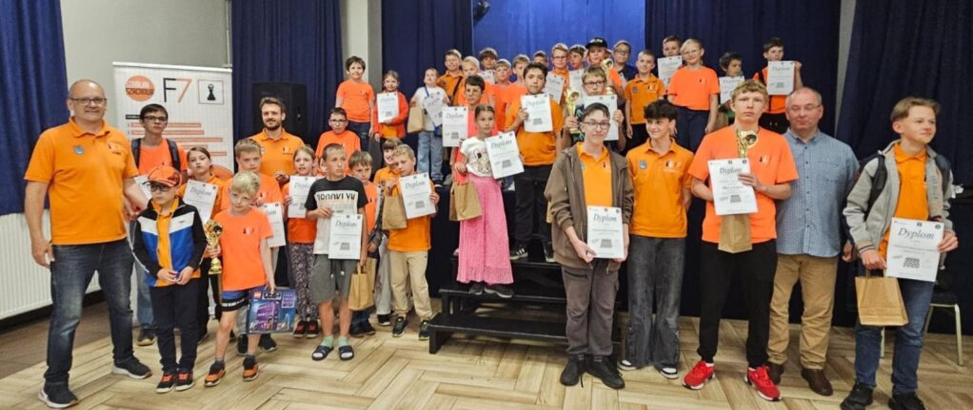 Grupa dzieci w pomarańczowych koszulka pozuje do zdjęcia z dyplomami w rękach