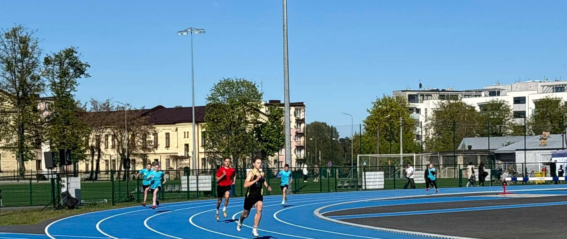 Sześcioro uczniów ubranych w stroje sportowe biegnie po stadionie lekkoatletycznym.