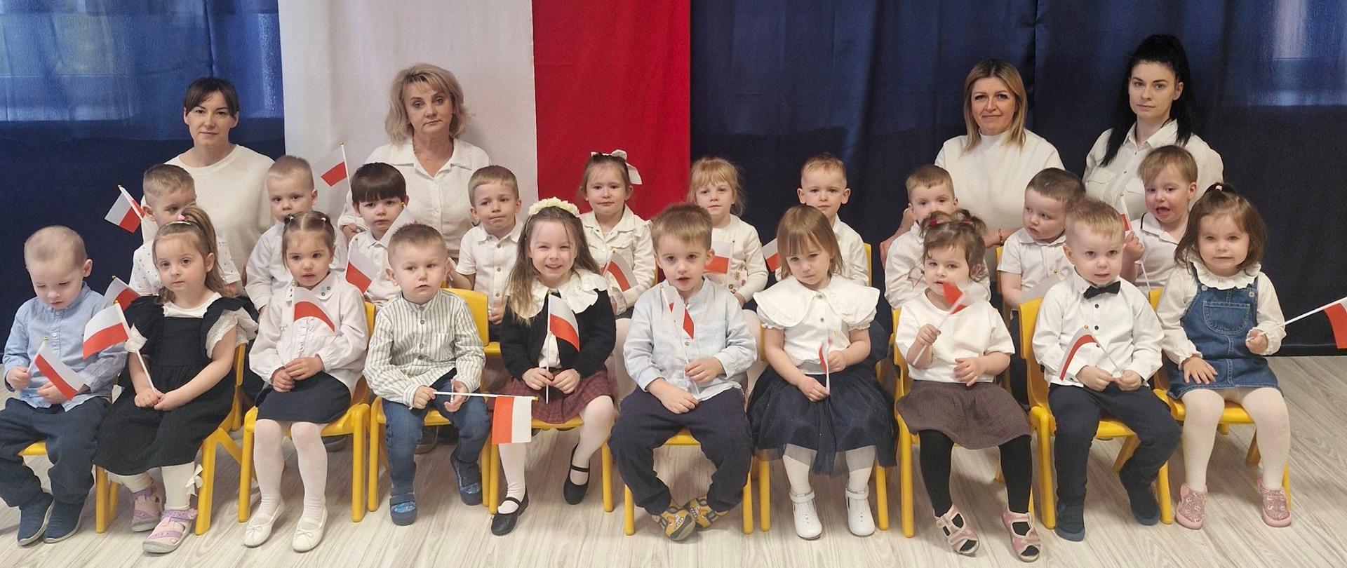Na zdjęciu są dzieci oraz panie z grupy Delfinki. Wszyscy są odświętnie ubrani. W tle widnieje tematyczna dekoracja.