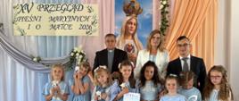 Grupa dziewczynek w niebieskich sukienkach i kompletach stoi na scenie obok kobiety w białym żakiecie i dwóch mężczyzn. Dziewczynka w środku trzyma pamiątkowy dyplom, a pozostałe dzieci trzymają maskotki.