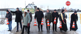 Dziewięć osób stoi w rzędzie na nowo wybudowanej drodze w Paproci Dużej. Zamiast tradycyjnego przecinania wstęgi, uczestnicy trzymają pomarańczowo-białe pachołki drogowe, które symbolizują bariery pokonane podczas realizacji inwestycji. Wśród obecnych są przedstawiciele władz powiatu, gminy oraz duchowieństwo. W tle widoczny jest znak "STOP" oraz maszyny budowlane.