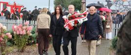 Trzyosobowa delegacja w eleganckich strojach niesie wieniec w barwach narodowych, udając się alejką pod pomnik.