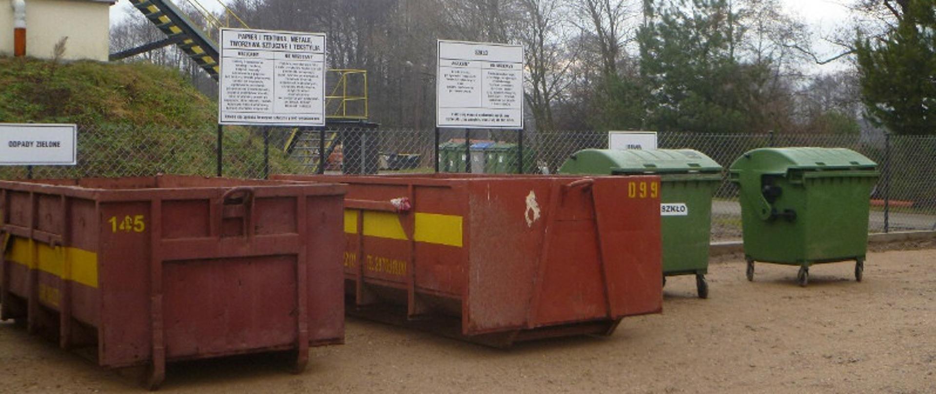 Dwa ciemno czerwone stalowe kontenery, dwa duże zielone pojemniki na śmieci na kołach, nad nimi stojące białe tablice