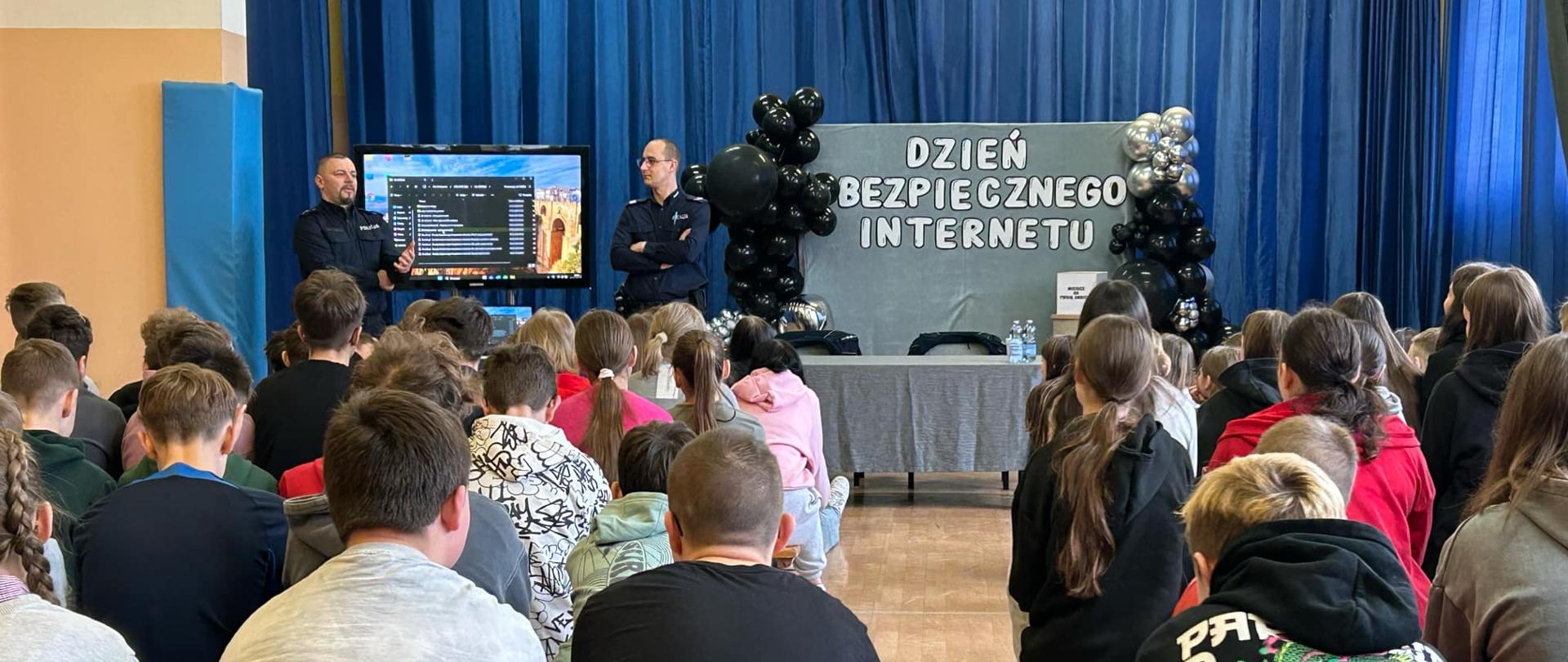 Na dużej sali dwoje policjantów w mundurach stoi obok telewizora. Obok nich stoi tablica z napisem Dzień bezpiecznego internetu. Przed nimi siedzi duża grupa młodzieży na krzesłach