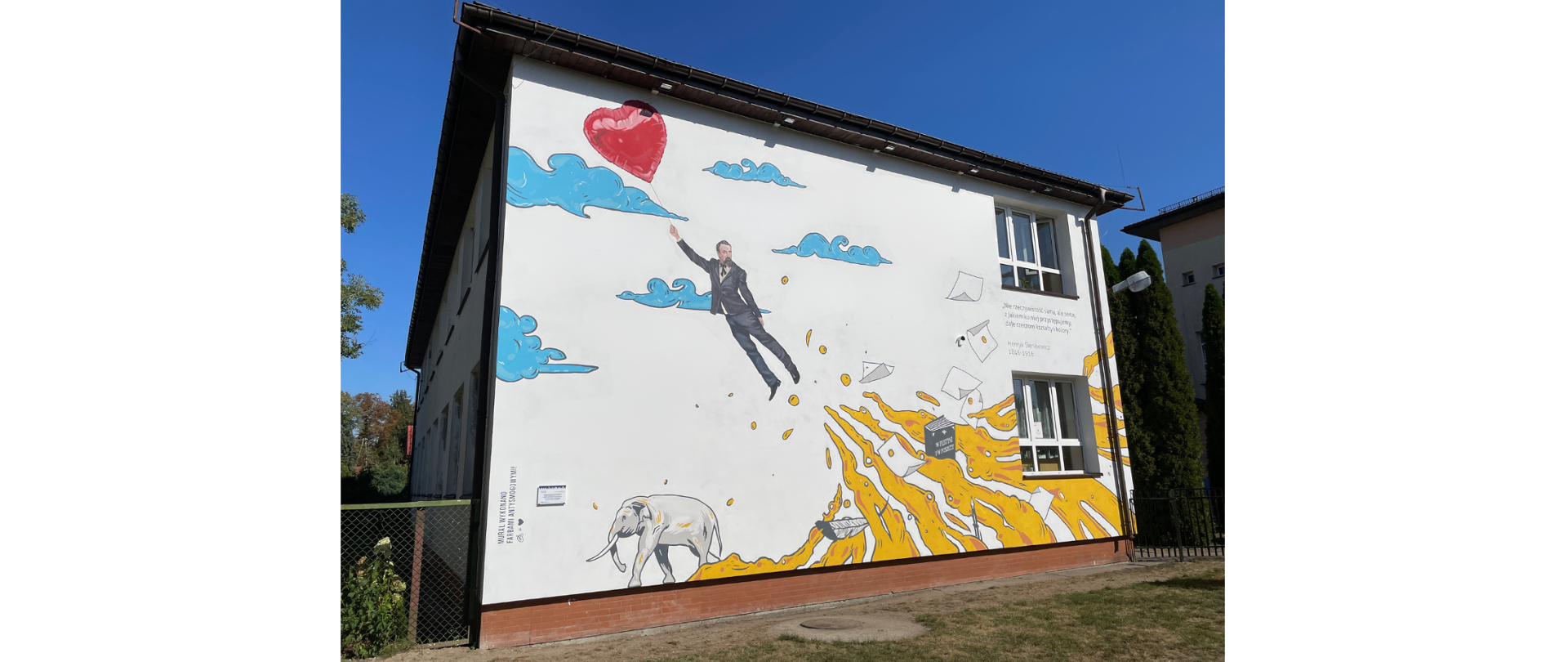 Budynek szkoły na którego ścianie namalowany został mural przedstawiający odlatującego Henryka Sienkiewicza, książki, chmury oraz słonia.