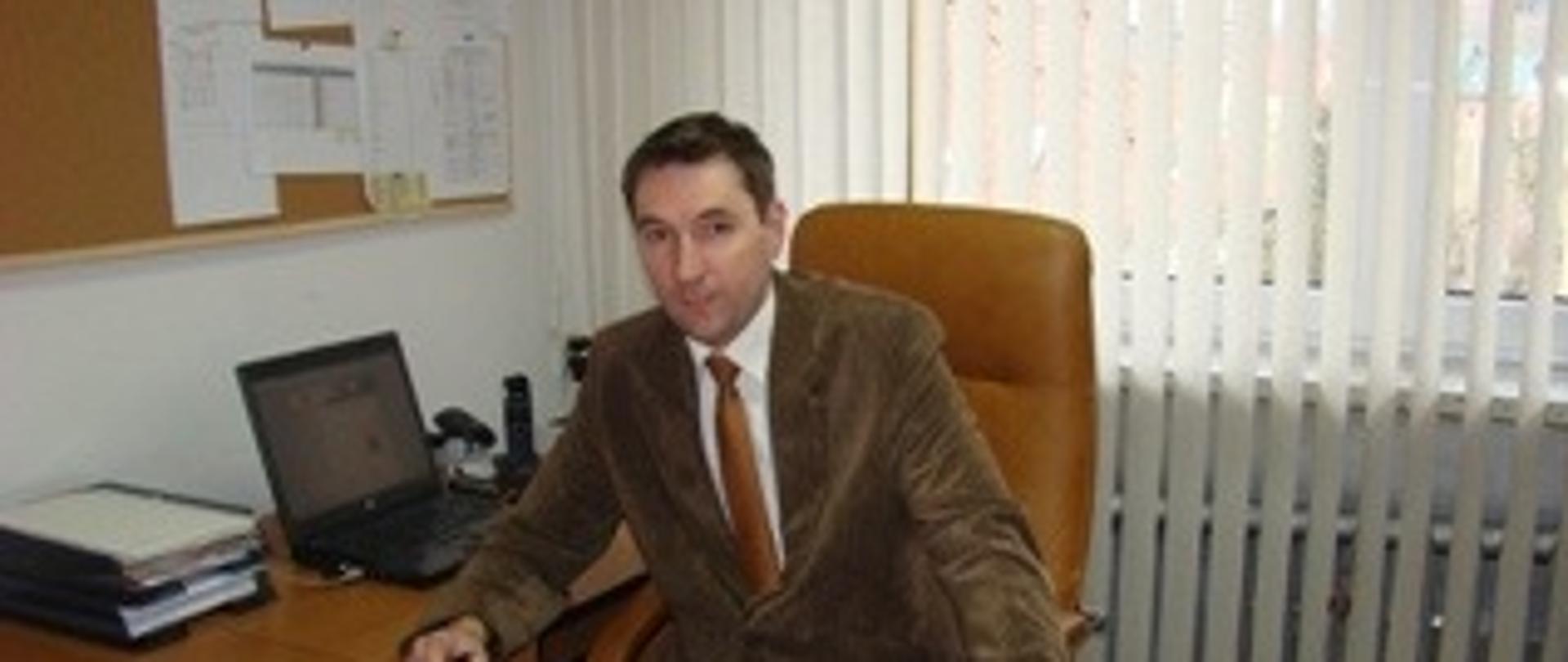 Zastępca Wójta Gminy Lubenia za biurkiem ręce na biurku brązowa marynarka w tle żaluzje okno i tablica korkowa