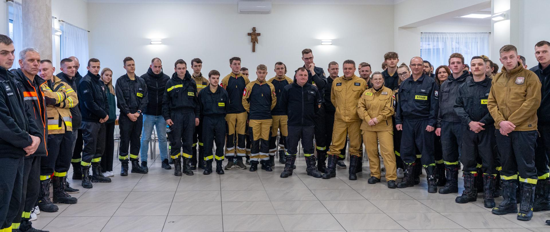 Grupowe zdjęcie druhów Ochotniczych Straży Pożarnych stojących w jasnej sali. Uczestnicy ubrani są w umundurowanie i odzież specjalistyczną OSP w kolorach czarnym i piaskowym, z elementami odblaskowymi. Strażacy ustawieni są w półokręgu, zwróceni w stronę aparatu. W tle widoczna jest jasna ściana z krzyżem oraz okna z firanami. Zdjęcie dokumentuje uroczyste spotkanie i wręczenie certyfikatów.