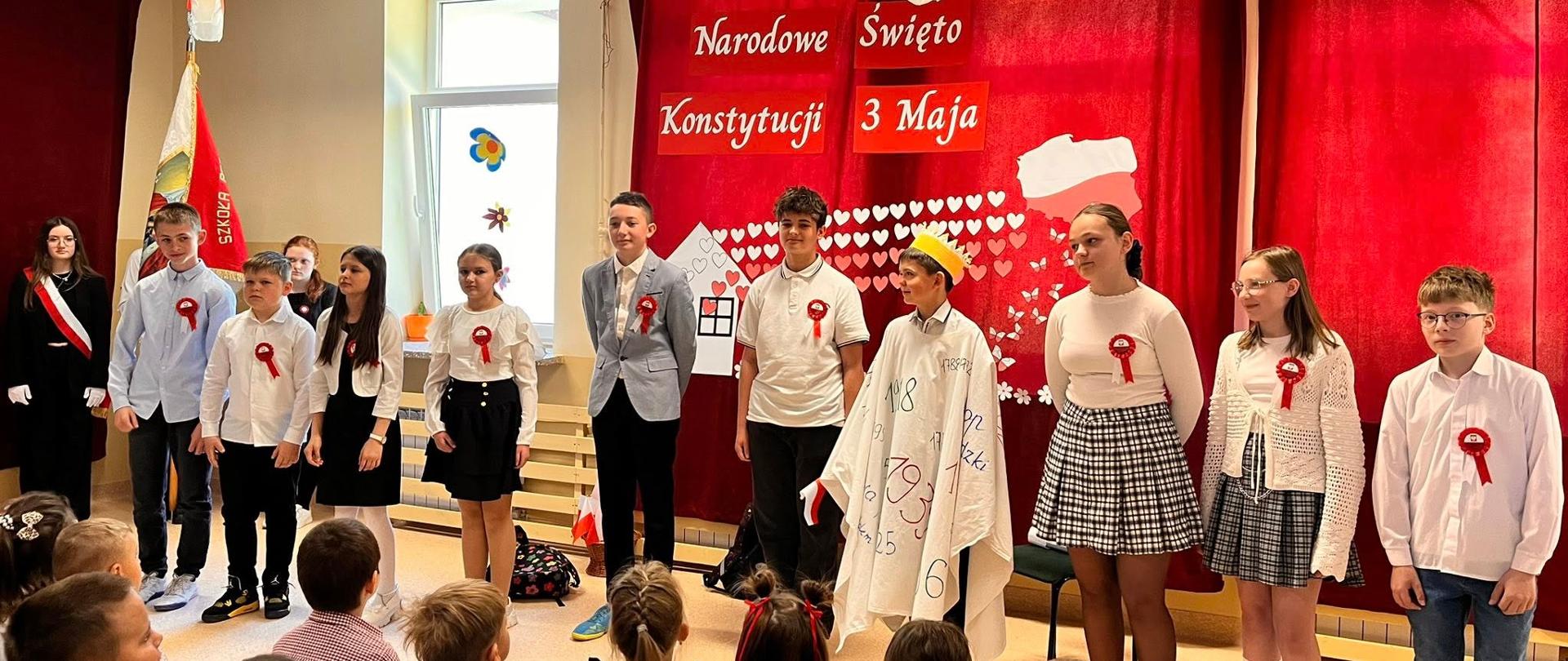 Uczniowie ubrani na galowo, z biało-czerwonymi kotylionami przypiętymi na piersi, występują na tle czerwonej kotary z biało-czerwonymi symbolami, uczeń w środku odgrywa króla