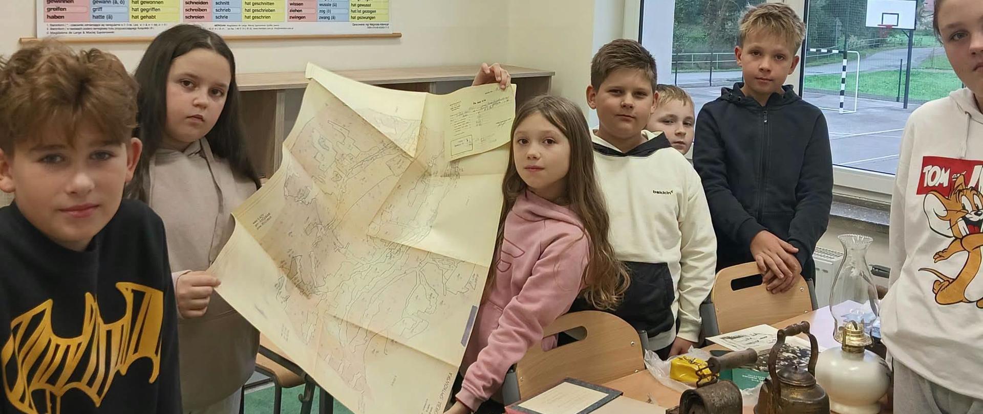 Na zdjęciu widać grupę dzieci w sali szkolnej. Kilkoro z nich stoi przy ławce i prezentuje dużą, rozłożoną mapę. Na stole znajdują się różne stare przedmioty, m.in. metalowe naczynia, lampy lub latarnie oraz dokumenty i zeszyty.
Na ścianie wisi duża tablica z niemieckimi formami czasowników nieregularnych i mieszanych. Przez okna widać boisko sportowe oraz drzewa. Zdjęcie wygląda na wykonane podczas lekcji lub zajęć związanych z historią, geografią albo dawnymi przedmiotami.