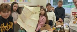 Na zdjęciu widać grupę dzieci w sali szkolnej. Kilkoro z nich stoi przy ławce i prezentuje dużą, rozłożoną mapę. Na stole znajdują się różne stare przedmioty, m.in. metalowe naczynia, lampy lub latarnie oraz dokumenty i zeszyty.
Na ścianie wisi duża tablica z niemieckimi formami czasowników nieregularnych i mieszanych. Przez okna widać boisko sportowe oraz drzewa. Zdjęcie wygląda na wykonane podczas lekcji lub zajęć związanych z historią, geografią albo dawnymi przedmiotami.