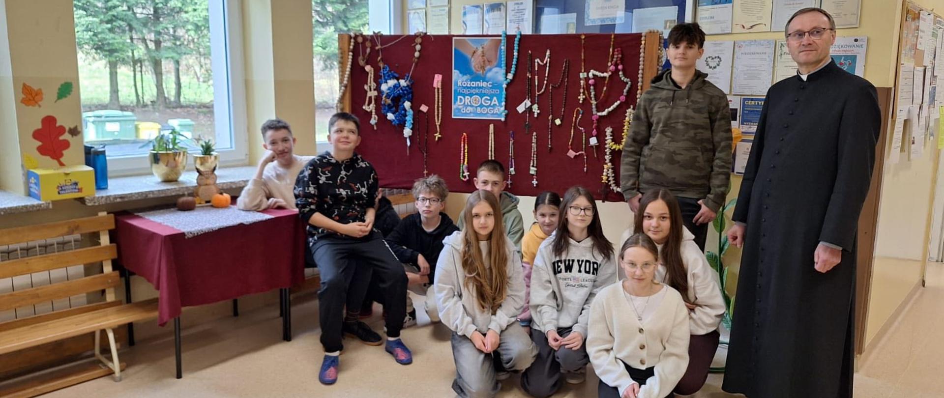 Nagrodzeni w konkursie uczniowie klasy 6 siedzą na korytarzu szkolnym, za nimi stojak, na którym powieszone są wykonane w konkursie różańce, po prawej stronie stoi Ksiądz