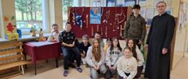 Nagrodzeni w konkursie uczniowie klasy 6 siedzą na korytarzu szkolnym, za nimi stojak, na którym powieszone są wykonane w konkursie różańce, po prawej stronie stoi Ksiądz