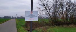 Zdjęcie przedstawia wiejską drogę z wysokim słupem po jej stronie. Na szczycie słupa zamontowane są dwa panele słoneczne, a poniżej znajduje się tablica informacyjna z tekstem w języku polskim oraz emblematem u góry. W tle widoczne są zielone pola, drzewa i pochmurne niebo.